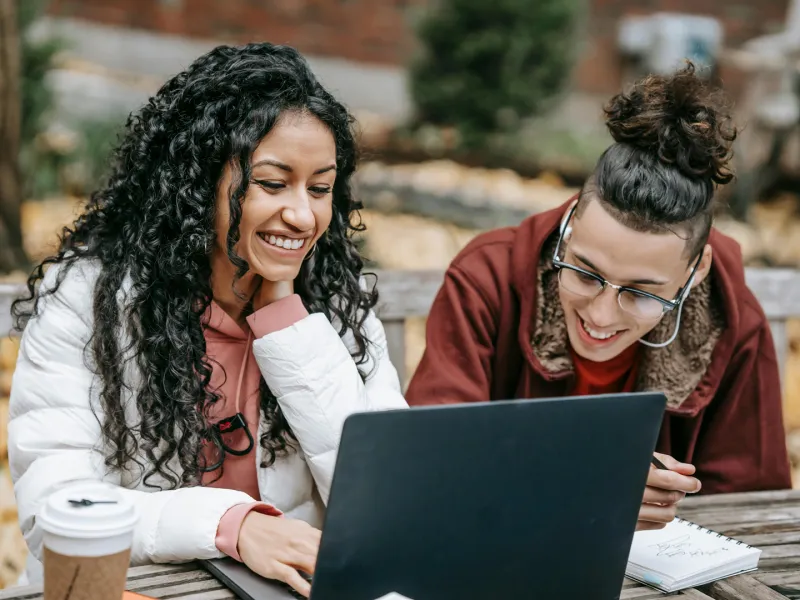 Zwei Studierende sitzen draußen auf einer Bank und schauen gemeinsam auf einen Laptop, umgeben von Pflanzen