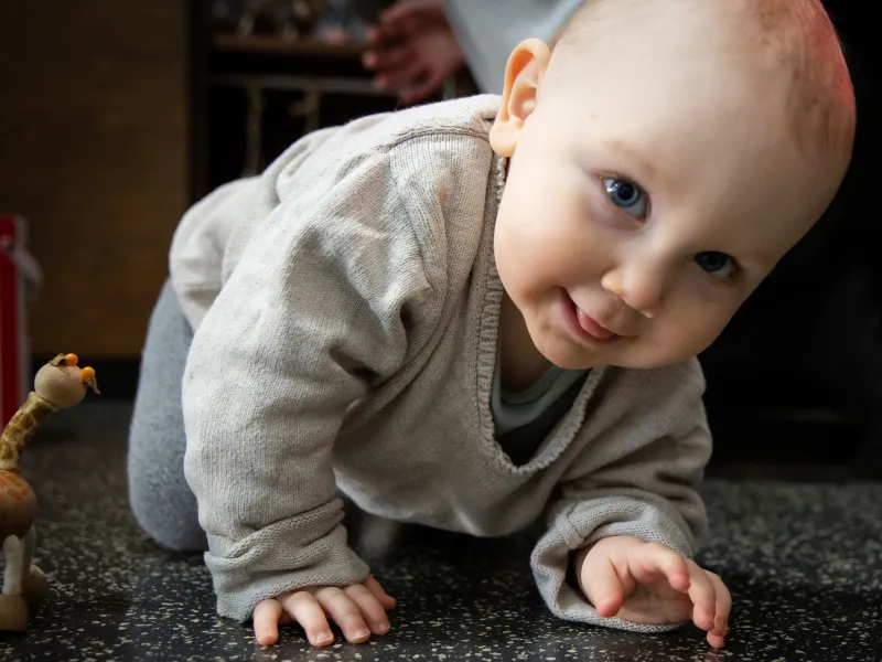 Kleinkind krabbelt auf einem dunklen, strukturierten Boden in einer Spielecke mit einem kleinen Spielzeugtier in der Nähe.