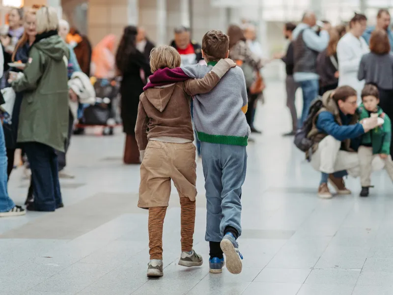 Zwei Kinder gehen Arm in Arm durch eine belebte Halle mit mehreren Menschen im Hintergrund.