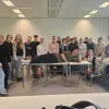 Eine Gruppe Studierender steht und sitzt in einem Klassenraum mit weißen Wänden und Deckenleuchten; Robert Fischbach liegt auf zwei Tischen in der Mitte.