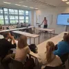 Ruben Fukkink steht vor einem Bildschirm und spricht in einem Klassenraum mit mehreren Zuhörenden, die an Tischen sitzen und ihm aufmerksam folgen.