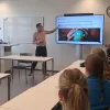 Ruben Fukkink steht vor einem Bildschirm und spricht in einem Klassenraum mit mehreren Zuhörenden, die an Tischen sitzen und ihm aufmerksam folgen.